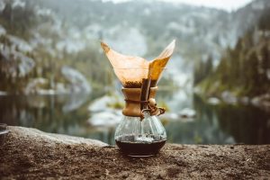Chemex, a design coffee maker on the ground in front of a forest lake.